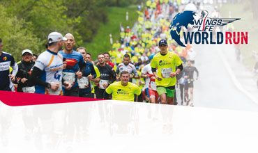 Eine große Gruppe unterschiedlicher Läufer, die am Wings for Life World Run teilnehmen, läuft eine malerische, von Bäumen gesäumte Straße entlang. Das Veranstaltungslogo ist in der oberen rechten Ecke zu sehen. Einige Teilnehmer tragen leuchtend gelbe Shirts.