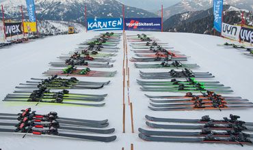 Reihenweise Skier sind auf einem schneebedeckten Berghang aufgereiht. Bunte Skier, darunter grüne, rote und gelbe, sind ordentlich angeordnet. Im Hintergrund zeigen Banner Logos und schneebedeckte Berge sind unter einem bewölkten Himmel sichtbar.