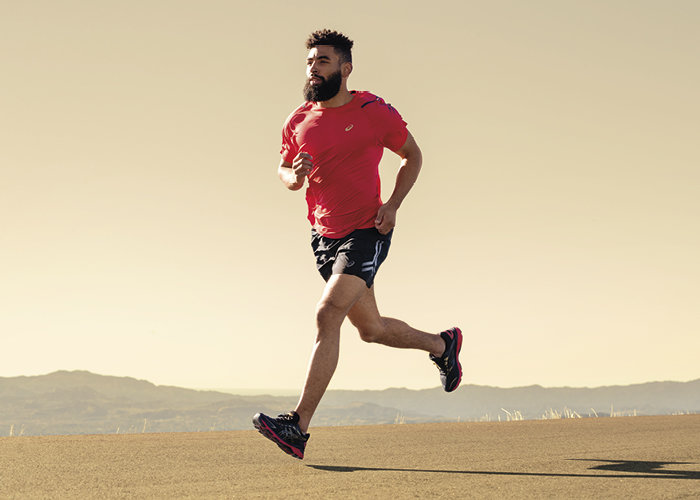 Ein Mann mit Bart läuft auf einer asphaltierten Straße. Er trägt ein rotes Hemd, schwarze Shorts und Laufschuhe. Im Hintergrund sind ein klarer Himmel und in der Ferne Berge zu sehen.