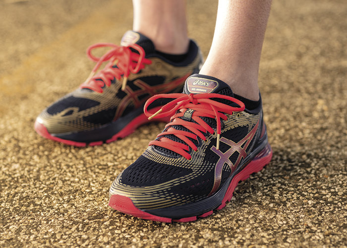 Nahaufnahme einer Person, die Sportschuhe mit einem schwarz-roten Design trägt und auf strukturiertem Boden steht. Die Schuhe haben rote Schnürsenkel und ein Logo an der Seite. Der Hintergrund ist leicht verschwommen.