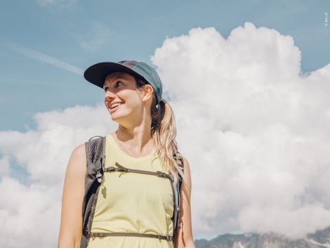 Lächelnde Frau vor einem Hintergrund aus flauschigen Wolken, mit Mütze und Rucksack. Sie trägt ein ärmelloses hellgelbes Oberteil und helles Tageslicht erhellt die Szene. In der Ferne sind unter dem klaren blauen Himmel schwach Berge zu erkennen.