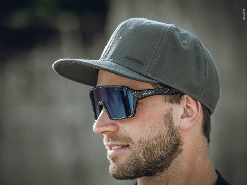 Eine Person mit einer grünen Baseballkappe und dunkler Sonnenbrille blickt zur Seite. Die Person hat einen Bart und der Hintergrund ist unscharf, wodurch ihr Profil hervorgehoben wird.