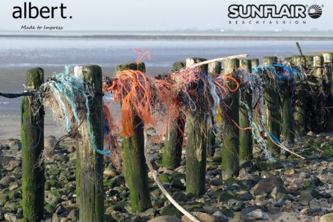 Verwitterte Holzpfosten an einem felsigen Strand sind mit verhedderten, bunten Fischernetzen und Seilen geschmückt. Im Hintergrund sind ein ruhiges Meer und eine entfernte Küstenlinie zu sehen. In den oberen Ecken erscheinen die Logos für „albert.“ und „SUNFLAIR Beachfashion“.