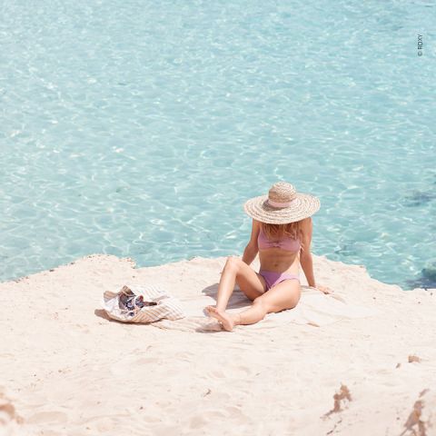 Eine Person in einem rosa Badeanzug und einem breitkrempigen Strohhut liegt an einem Sandstrand neben klarem, türkisfarbenem Wasser. Eine gestreifte Tasche und eine Sonnenbrille liegen in der Nähe im Sand.