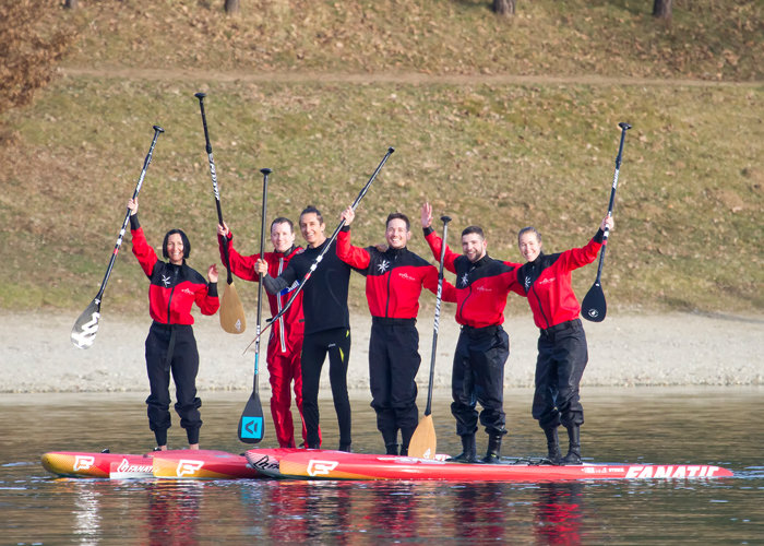 Sechs Personen in roten und schwarzen Neoprenanzügen stehen auf zwei Paddleboards auf einem ruhigen Gewässer. Sie lächeln, halten die Paddel aufrecht und strecken ihre Arme freudig in die Höhe. Im Hintergrund ist eine Grasfläche mit vereinzelten Bäumen zu sehen.