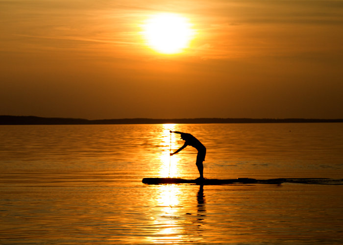 Eine Person paddelt bei Sonnenuntergang auf ruhigem Wasser. Die Silhouette ist vor dem goldenen Himmel zu sehen, wobei sich die Sonne auf der Wasseroberfläche spiegelt.