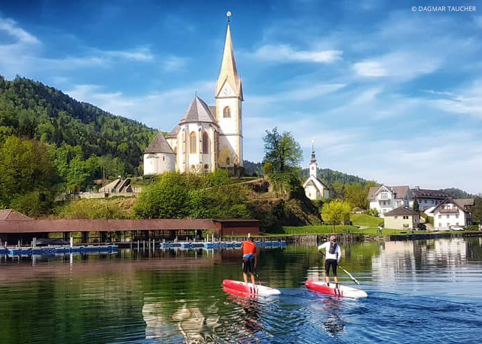 Zwei Personen paddeln auf einem ruhigen See und blicken auf ein malerisches Bergdorf mit einer markanten Kirche mit einem hohen Turm. Der Hintergrund ist eine Mischung aus üppigen grünen Hügeln und einem klaren blauen Himmel.