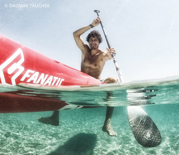 Ein Mann ohne Hemd sitzt auf einem roten Paddleboard in klarem, seichtem Wasser und hält in einer Hand ein Paddel. Das Foto ist teilweise unter Wasser und zeigt das Board und den Sandboden. Der Text auf dem Board lautet „Fanatic“.