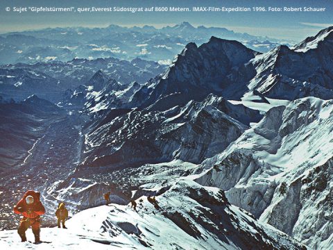 Bergsteiger überqueren einen schneebedeckten Grat nahe dem Gipfel des Mount Everest. Die Landschaft ist weitläufig, mit schroffen Gipfeln vor einem klaren Himmel. Die Bergsteiger stehen mit Abstand entlang des Pfades, einige tragen helle Jacken. In der oberen linken Ecke ist Text zu sehen.