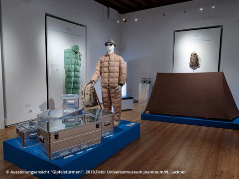 Museumsausstellung mit einer Bergsteigerausstellung mit Schaufensterpuppen in Pufferjacken, Schutzbrillen, Hüten und Rucksäcken. In der Nähe ist ein braunes Zelt aufgebaut. Metallkisten mit Ausrüstung sind auf blauen Plattformen angeordnet.