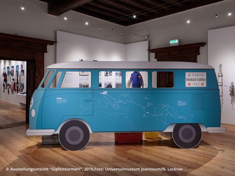 Ausstellungsstück eines blauen Retro-Vans aus den 1960er Jahren in einem Museumsraum, auf dessen Seite eine Weltkarte abgebildet ist. Umgeben von Exponaten mit Kleidung und Bergsteigerausrüstung.