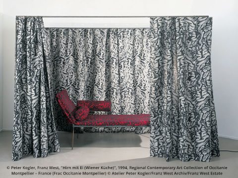 Eine Rauminstallation mit einer rot-schwarzen Chaiselongue mit abstrakten Mustern vor einem Hintergrund aus passenden gemusterten Vorhängen, die an einem Rahmen hängen. Das Design schafft eine visuell komplexe und immersive Umgebung.