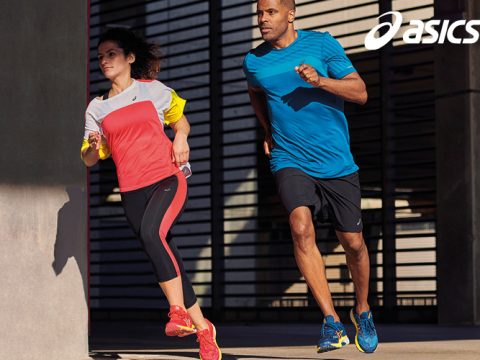 Zwei Personen joggen draußen in ASICS-Sportbekleidung. Die Frau trägt ein rot-weißes Outfit mit roten Schuhen, während der Mann ein blaues Hemd und schwarze Shorts mit blauen Schuhen trägt. Sie laufen an einem modernen Gebäude vorbei. In der Ecke ist das ASICS-Logo zu sehen.
