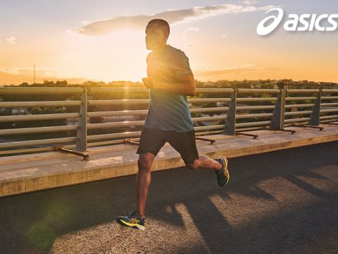 Eine Person läuft bei Sonnenuntergang über eine Brücke. Sie trägt ein blaues Shirt, schwarze Shorts und Laufschuhe. Im Hintergrund geht die Sonne unter und wirft einen goldenen Schein auf die Brücke. In der oberen rechten Ecke ist das ASICS-Logo zu sehen.