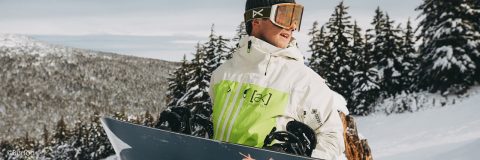 Eine Person in einer grün-weißen Jacke steht mit einem Snowboard in der Hand auf einem schneebedeckten Berghang. Sie trägt eine Schutzbrille und eine Mütze. Im Hintergrund sind schneebedeckte Bäume zu sehen.