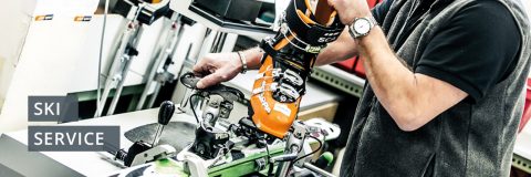 Eine Person passt in einer Werkstatt einen Skischuh an einem Ski an. Der Schuh ist orange und schwarz, der Ski ist grün und weiß. In der Nähe steht ein Schild mit der Aufschrift „SKISERVICE“. Die Person trägt eine Uhr und eine Weste.