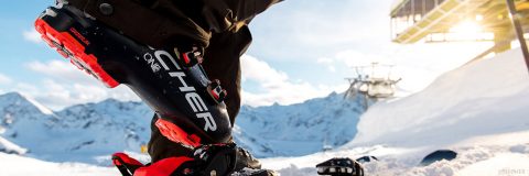 Nahaufnahme des Skischuhs eines Skifahrers, der in einer Skibindung in einer verschneiten Berglandschaft befestigt ist. Im Hintergrund sind die schneebedeckten Gipfel und ein Skilift unter einem klaren blauen Himmel zu sehen.