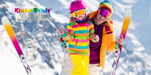 Ein Erwachsener und ein Kind in farbenfroher Skikleidung, einschließlich Helm und Schutzbrille, stehen glücklich mit Skiern auf einem schneebedeckten Berg. Sie lächeln und im Hintergrund ist eine verschwommene Schneelandschaft mit Bergen zu sehen.