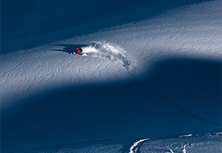 Ein Skifahrer in einer roten Jacke fährt durch den tiefen Schnee an einem Berghang und hinterlässt eine weiße Gischtspur. Die Szene wurde von oben aufgenommen, wodurch die kontrastierenden Texturen von Schnee und Schatten hervorgehoben werden.