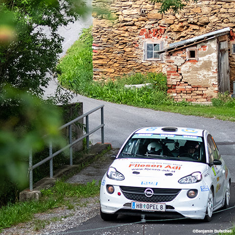Ein Rallyeauto mit Sponsorenlogos fährt eine Kurve auf einer Landstraße, die von einer Steinmauer und Grünflächen gesäumt ist. Im Hintergrund ist ein altes Gebäude aus Backstein und Stein mit verwittertem Aussehen zu sehen.