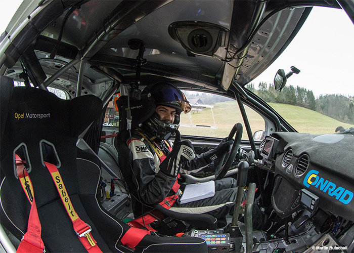 Ein Fahrer in Rennkleidung sitzt in einem Rallyeauto, hält mit einer Hand das Lenkrad und streckt mit der anderen den Daumen nach oben. Im Innenraum befinden sich Rennsitze und -ausrüstung. Draußen sind teilweise grasbewachsene Hügel und Bäume sichtbar.