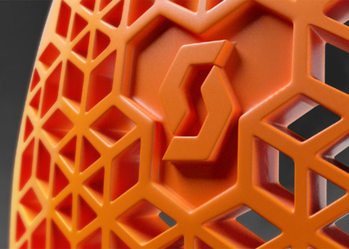 Nahaufnahme eines orangefarbenen Objekts mit einem geometrischen, sechseckigen Gittermuster und einem stilisierten Logo in der Mitte. Der Hintergrund ist unscharf.