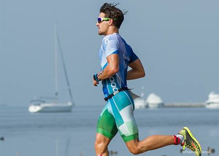 Eine Person in einem blau-grünen Triathlonanzug und mit Sonnenbrille läuft an einer Uferpromenade entlang, im Hintergrund sind Segelboote und ein ruhiges Meer zu sehen.