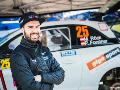 Mann in schwarzer Jacke und Mütze steht lächelnd mit verschränkten Armen vor einem weißen Rallyeauto mit der Nummer 25 und „M. Röck, P. Forstner“ auf der Scheibe. Das Auto steht unter einem Vordach.