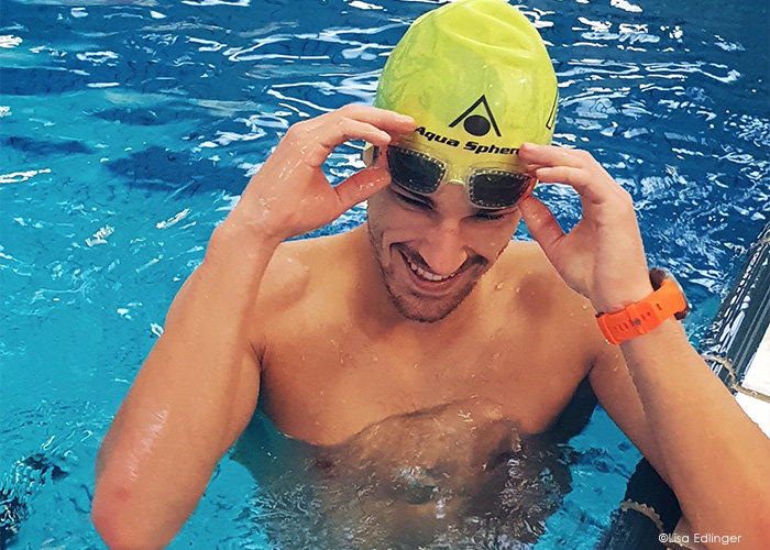 Ein Schwimmer mit limettengrüner Badekappe und orangefarbener Armbanduhr rückt seine Schwimmbrille zurecht, während er in einem Pool steht. Er lächelt und das Wasser um ihn herum ist blau.