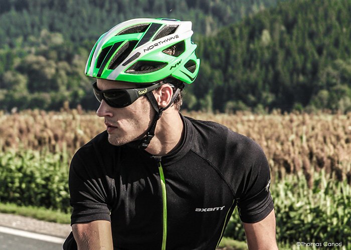 Ein Radfahrer mit einem schwarz-grünen Helm, einer schwarzen Sonnenbrille und einem schwarzen Radtrikot fährt auf einer Straße. Im Hintergrund sind Felder und dichte grüne Bäume zu sehen.