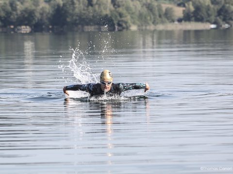 Eine Person in schwarzem Neoprenanzug und Schwimmbrille schwimmt im offenen Wasser und führt den Schmetterlingsstil aus. Um sie herum sind Spritzer zu sehen. Bäume säumen das entfernte Ufer unter einem klaren Himmel.
