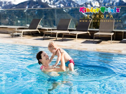 Eine Frau hält ein lächelndes Kind in einem Swimmingpool. Im Hintergrund sind Berge zu sehen. Auf dem Deck sind Sonnenliegen aufgereiht. In der oberen rechten Ecke steht der Schriftzug „Kinderhotels Europa“.