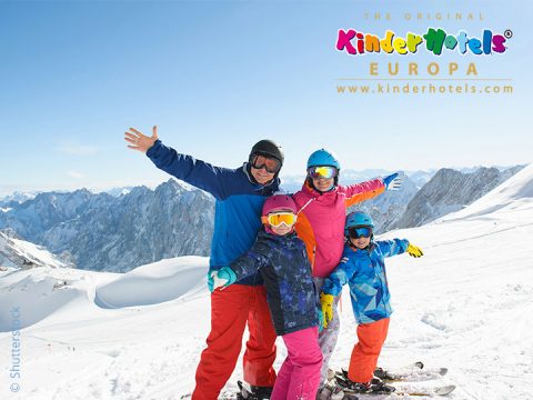 Eine vierköpfige Familie in Skiausrüstung posiert glücklich auf einem schneebedeckten Berg. Der Hintergrund zeigt einen klaren blauen Himmel und entfernte Berggipfel. Das Bild ist mit dem Logo von Kinderhotels Europa versehen.