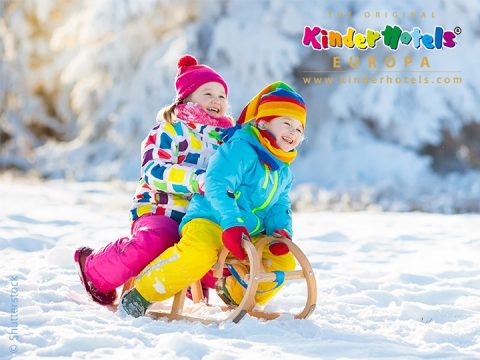 Zwei Kinder, eingepackt in bunte Winterkleidung, rodeln einen verschneiten Hang hinunter. Sie sitzen auf einem Holzschlitten und lachen vor Freude. Im Hintergrund sind schneebedeckte Bäume zu sehen. Das Bild ist oben rechts mit „Kinderhotels Europa“ beschriftet.