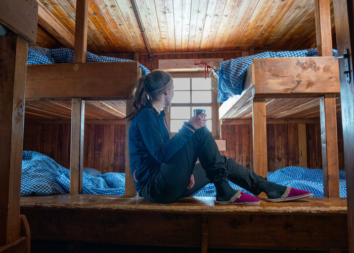 Eine Person sitzt auf einem Holzboden in einer gemütlichen Hütte und hält eine Tasse in der Hand. Sie trägt dunkle Kleidung und blickt zu einem kleinen Fenster. Sie ist von Etagenbetten mit blau karierter Bettwäsche umgeben und die Hütte hat eine warme, hölzerne Inneneinrichtung.
