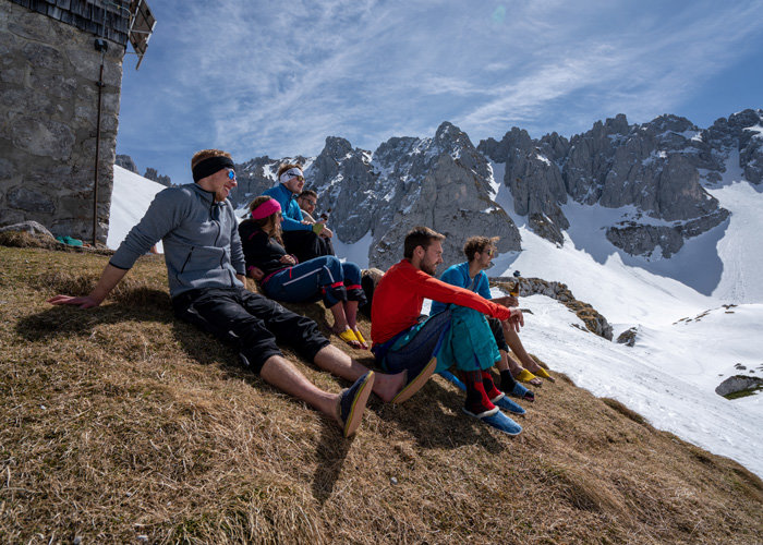 Eine Gruppe von sechs Personen in farbenfroher Wanderkleidung sitzt auf einem Grashang und blickt auf eine schneebedeckte Bergkette unter blauem Himmel. Auf der linken Seite ist teilweise ein rustikales Steingebäude zu sehen. Sie wirken entspannt und genießen die Aussicht.