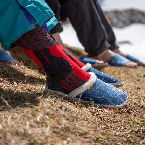 Nahaufnahme mehrerer Personen, die auf Gras sitzen und blaue Slipper mit weißem Fellfutter tragen. Eine Person trägt bunte rote und schwarze Socken und blaue Hosen. Die Szene spielt im Freien, im Hintergrund sind teilweise die Beine und Füße der anderen Personen zu sehen.