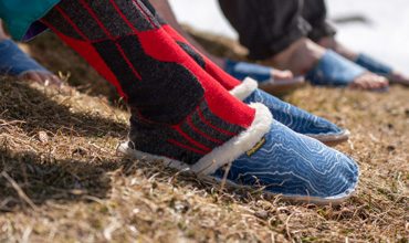 Nahaufnahme einer Person mit roten und schwarzen Socken und blauen Hausschuhen, die auf trockenem Gras steht. Im Hintergrund ist eine weitere Person in blauen Hausschuhen teilweise zu sehen. Die Hausschuhe haben ein Fleecefutter und ein weißes Wirbelmuster.