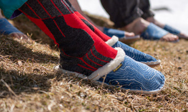 Nahaufnahme einer Person mit roten und schwarzen Socken und blauen Hausschuhen, die auf trockenem Gras steht. Im Hintergrund ist eine weitere Person in blauen Hausschuhen teilweise zu sehen. Die Hausschuhe haben ein Fleecefutter und ein weißes Wirbelmuster.