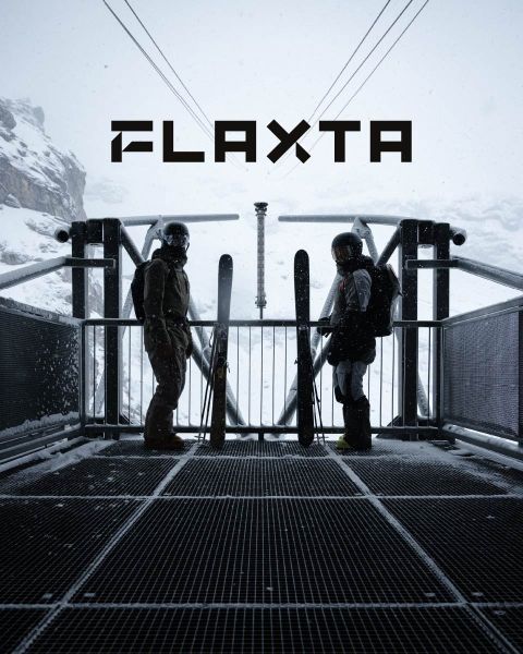 Zwei Skifahrer mit Helmen und Ausrüstung stehen auf einer schneebedeckten Metallplattform mit Blick auf eine schneebedeckte Berglandschaft. Sie halten Skier in den Händen und stehen sich gegenüber. Über ihnen prangt in fetten Buchstaben das Wort „FLAXTA“.