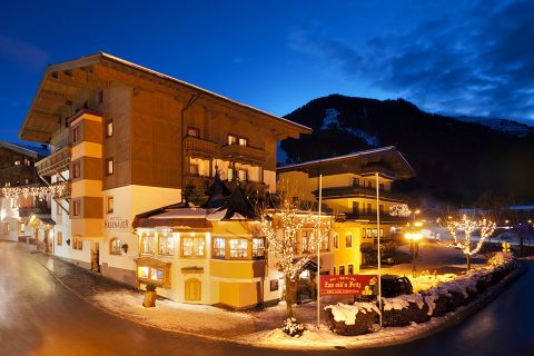 Charmante Szene eines Alpendorfs in der Abenddämmerung mit festlich beleuchteten traditionellen Gebäuden. Schnee bedeckt den Boden und die Berge in der Ferne bilden eine malerische Kulisse vor einem tiefblauen Himmel.