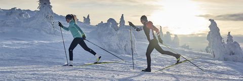 Zwei Personen laufen in einer verschneiten Landschaft unter einem bewölkten Himmel Skilanglauf. Sie tragen Wintersportkleidung und sind von schneebedeckten Bäumen umgeben. Die Sonne steht tief am Horizont und verbreitet einen warmen Schein.