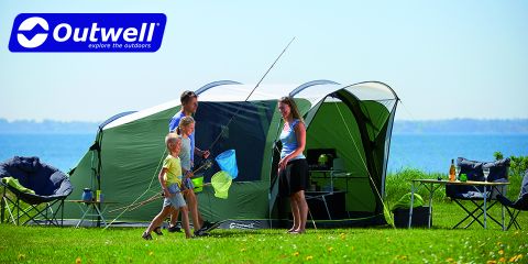 Eine vierköpfige Familie genießt einen sonnigen Tag beim Camping am Wasser. Zwei Kinder halten Fischernetze, während zwei Erwachsene daneben stehen. Ein großes grünes Zelt ist aufgebaut, umgeben von Campingstühlen und -ausrüstung. Das Outwell-Logo ist in der oberen linken Ecke zu sehen.