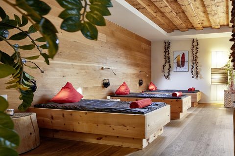 Ein gemütlicher Spa-Raum mit drei hölzernen Lounge-Stühlen, jeder mit einem grauen Polster und einem roten Kissen. Auf den Stühlen liegen gerollte rote Handtücher. Warme Holzwände und -decken, grüne Pflanzen und sanfte Beleuchtung verstärken die entspannende Atmosphäre.