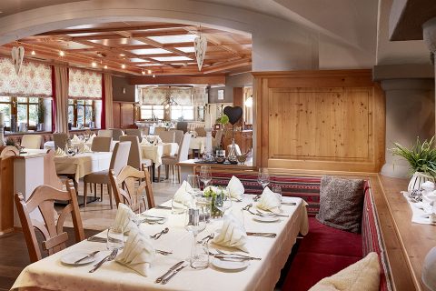 Ein gemütliches Restaurant-Interieur mit Holzakzenten und elegantem Dekor. Die Tische sind mit weißer Tischwäsche, gefalteten Servietten, Gläsern und Besteck gedeckt. Große Fenster lassen Tageslicht herein und gepolsterte Sitzgelegenheiten sorgen für eine angenehme Essatmosphäre.
