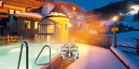 Beheizter Außenpool in einem verschneiten Resort mit Gebäuden im Hintergrund. Warmes Licht erhellt den Bereich und bildet einen Kontrast zum kühlen Abendhimmel. Im unteren Teil des Bildes ist das Wappen des Resorts zu sehen.