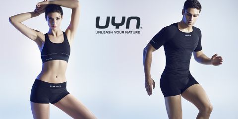 Eine Frau und ein Mann tragen schwarze Sportbekleidung mit dem „UYN“-Logo. Die Frau trägt einen Sport-BH und Shorts, der Mann ein figurbetontes Shirt und Shorts. Über ihnen steht der Text „UNLEASH YOUR NATURE“.