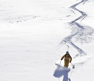 Ein Skifahrer in gelbem Outfit und Helm gleitet unter klarem Himmel einen schneebedeckten Hang hinunter und hinterlässt eine gewundene Spur im frischen Pulverschnee.