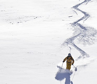 Ein Skifahrer in gelbem Outfit und Helm gleitet unter klarem Himmel einen schneebedeckten Hang hinunter und hinterlässt eine gewundene Spur im frischen Pulverschnee.