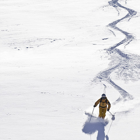 Ein Skifahrer in gelbem Outfit gleitet einen schneebedeckten Hang hinunter und hinterlässt eine gewundene Spur. Die Szene ist hell von unberührtem Schnee und der Skifahrer wirft einen Schatten auf die weiße Oberfläche.